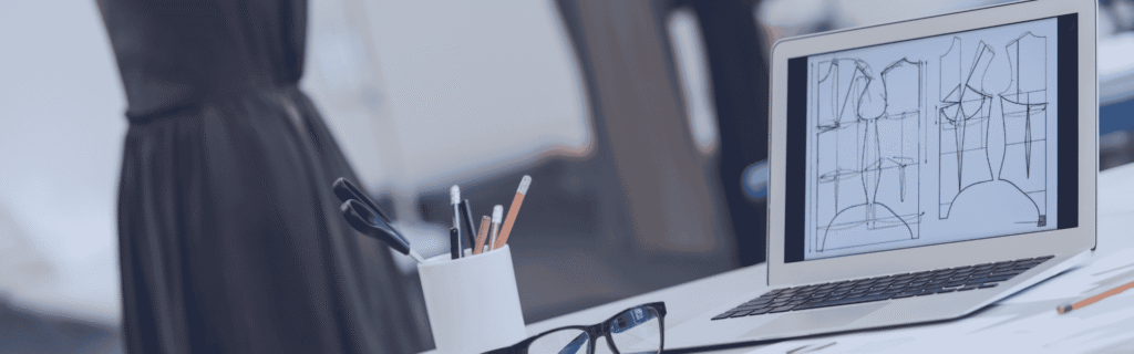 A desk with a laptop, sketches and eyeglasses on it, dress models on background