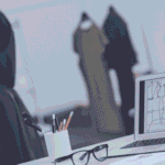 A desk with a laptop, sketches and eyeglasses on it, dress models on background