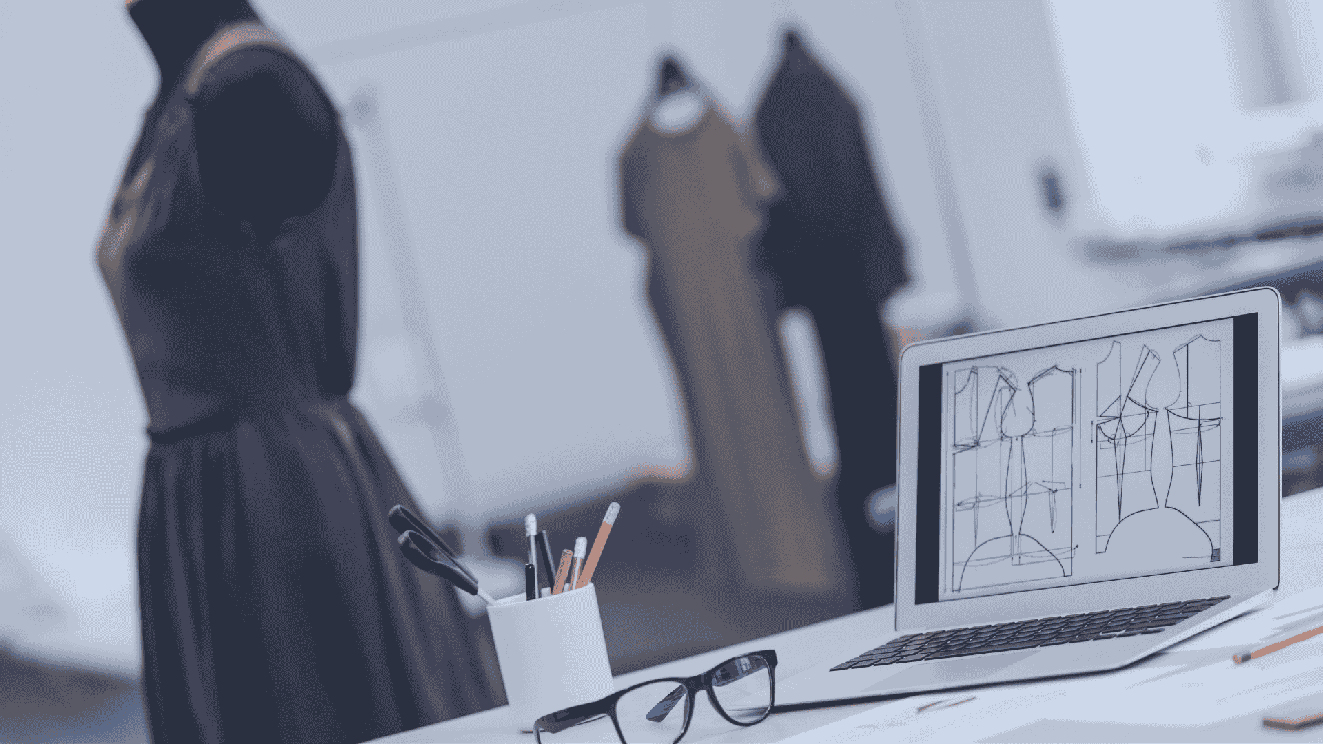 A desk with a laptop, sketches and eyeglasses on it, dress models on background